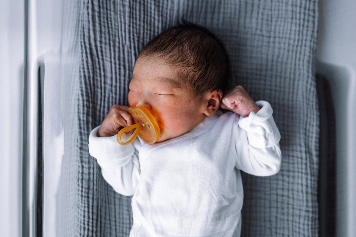 newborn sleeping in hospital bassinet with pacifier and arms up near face