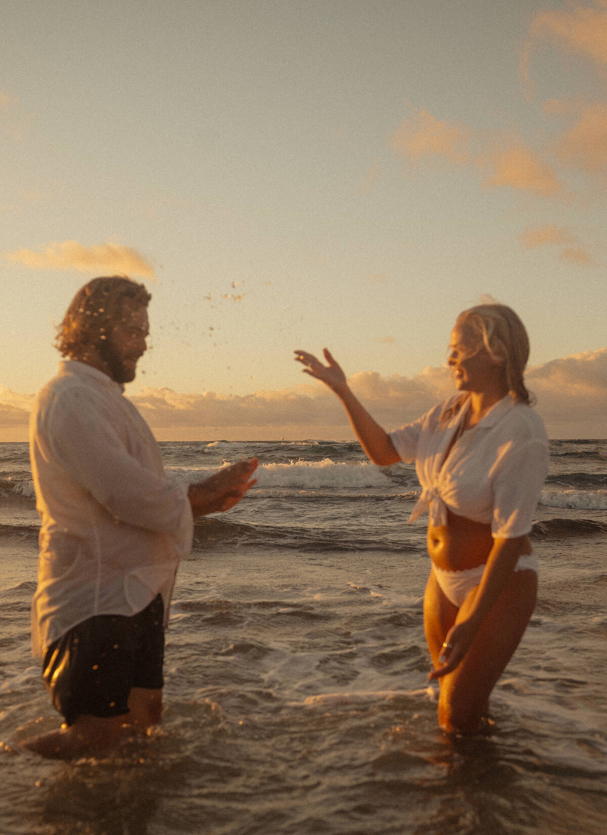 elopement couple playfully splash each other as they stand in the ocean and exchange their wedding vows among the sunset sky and waves