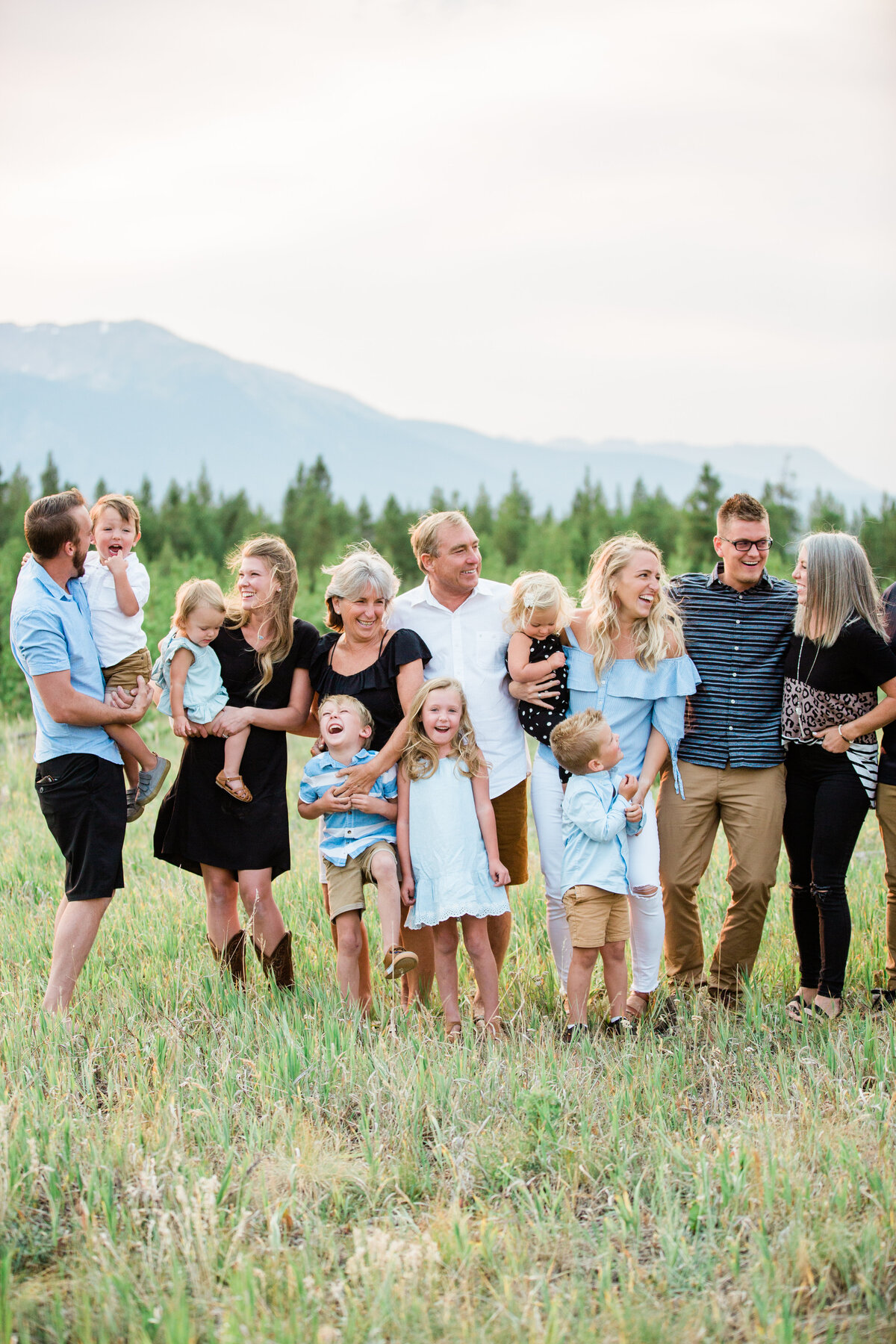 A large extended family are all laughing and smiling with one another in a mountain meadow in the summer.
