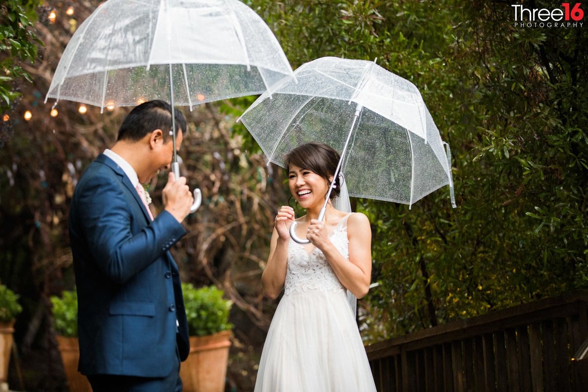 Bride and Groom share a laugh while holding umbrellas during rainfall