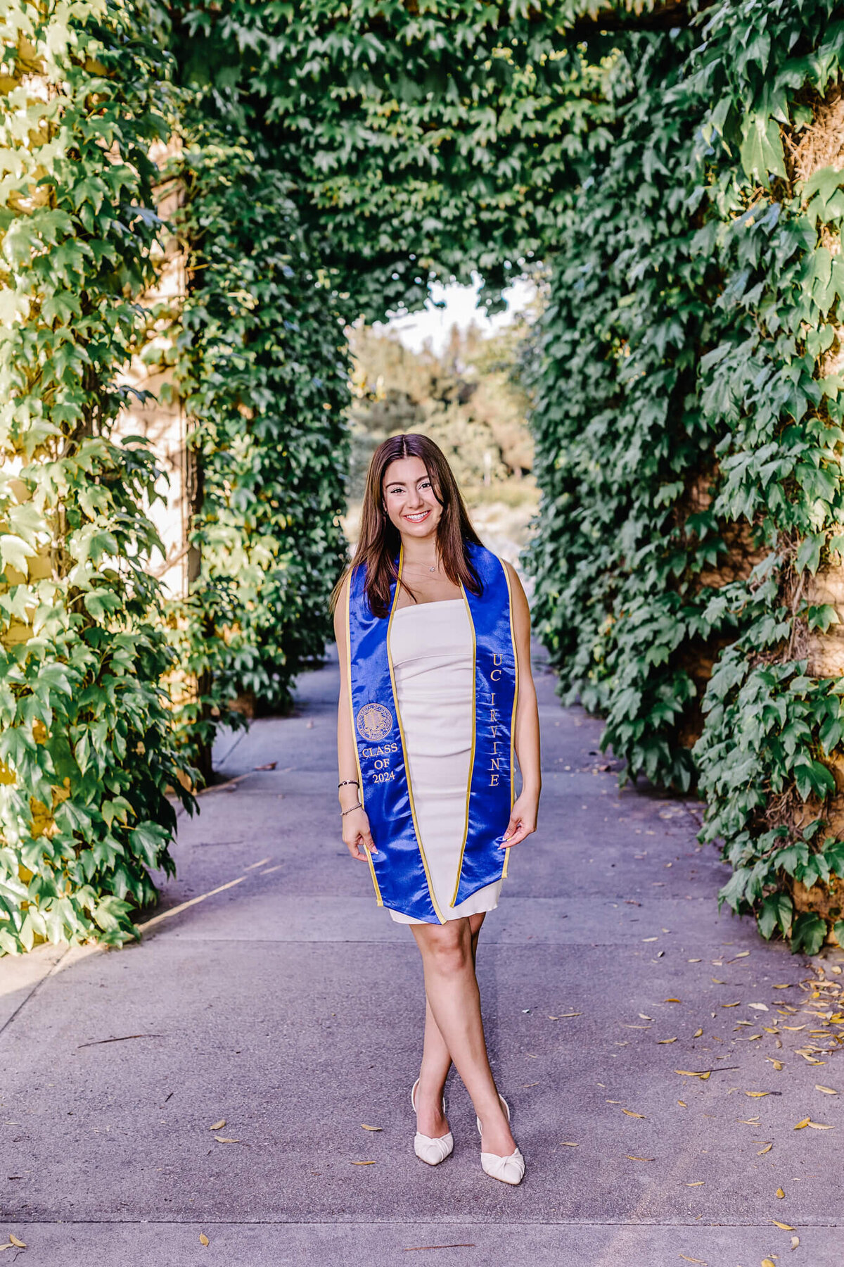 A young woman in a white dress and blue graduation stole stands on a pathway adorned with lush green ivy. She is smiling and posing confidently, with sunlight filtering through the leaves.