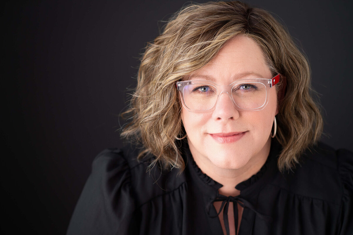 a woman in a black blouse and eyeglasses leaning forward and smiling softly.  Captured in-studio by Ottawa headshot photographer JEMMAN  Photography