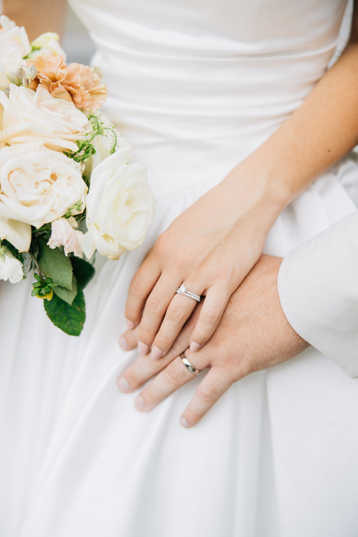 Bride & Groom hold hands with their wedding rings showing and bride holding a bouquet of soft peach roses