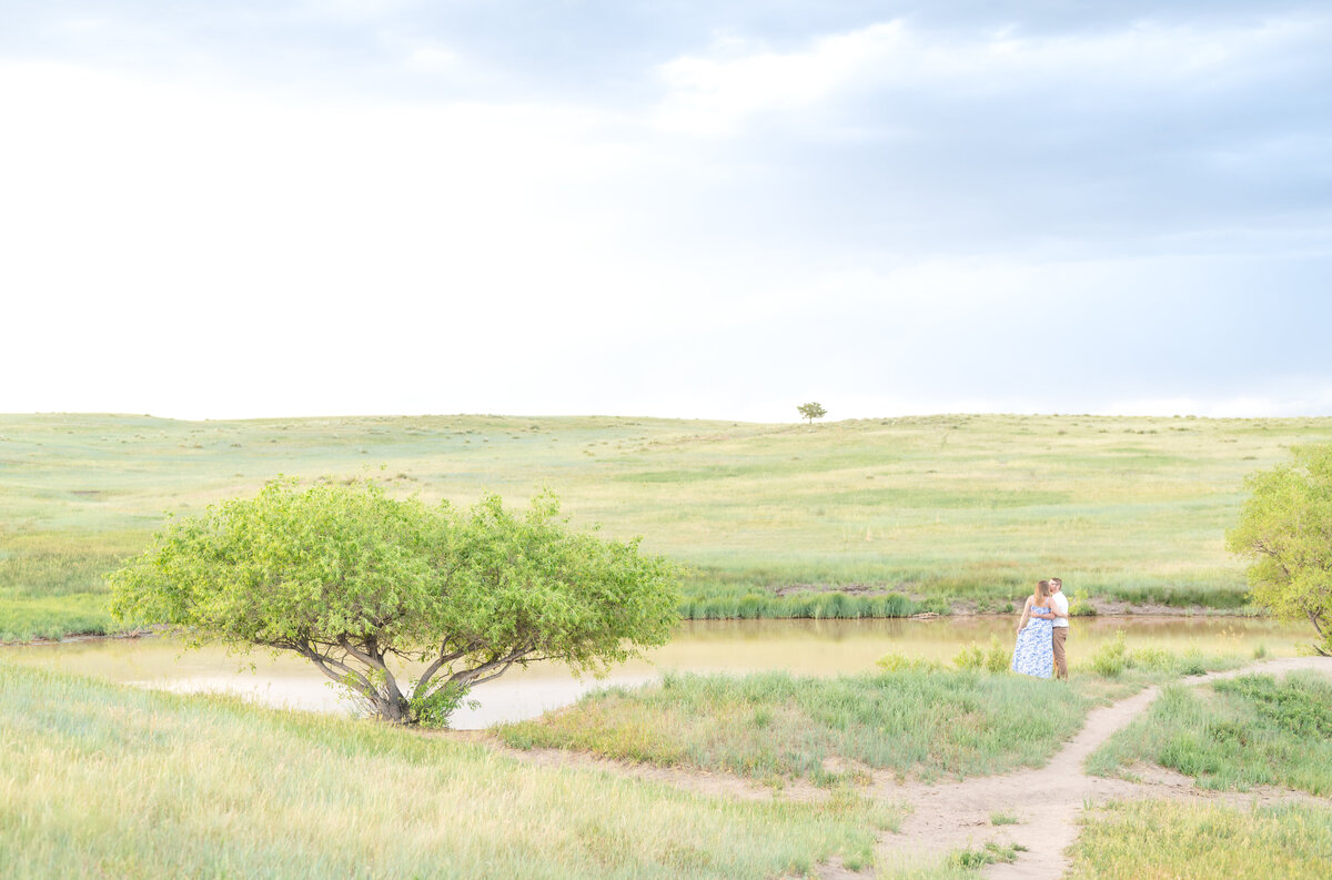 Wide landscape photo of couple hugging while standing near water in mountain meadow field looking out into the distance