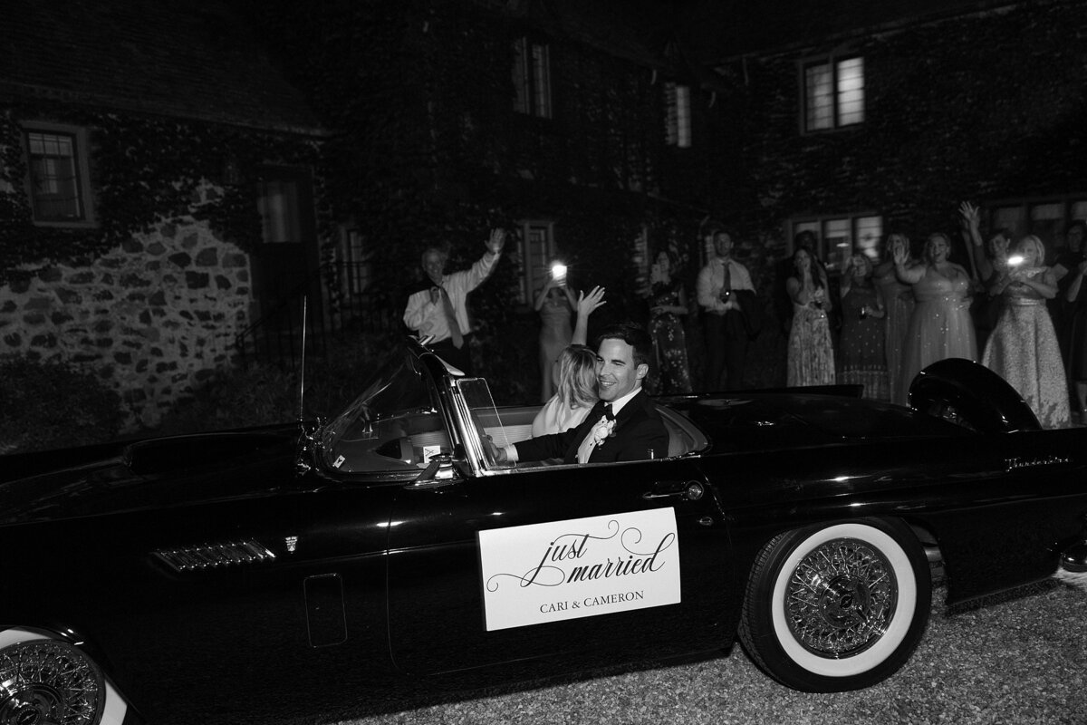 Bride and Groom leave wedding reception in vintage car as guests wave in the background.
