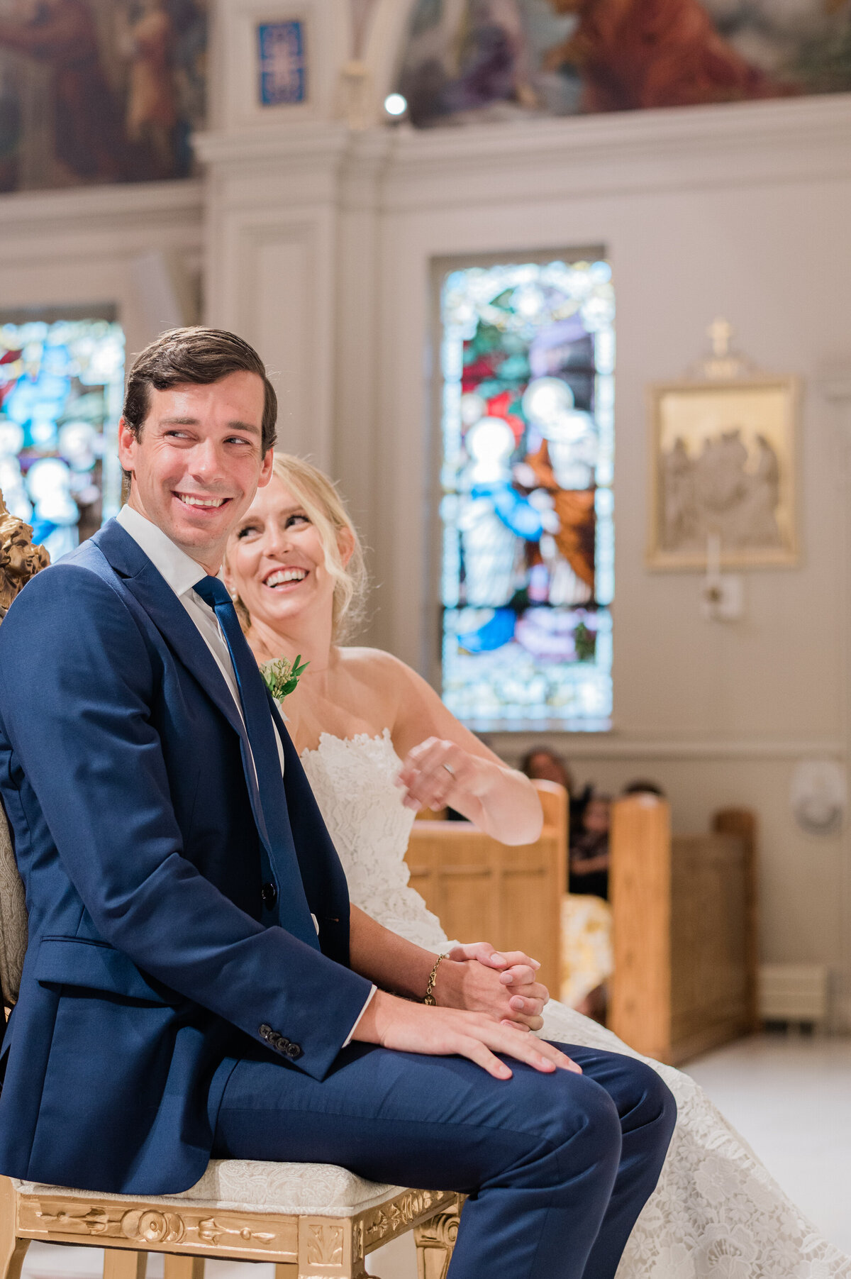 A joyful groom in a navy suit and a glowing bride in a white gown share a smile while sitting in the church.