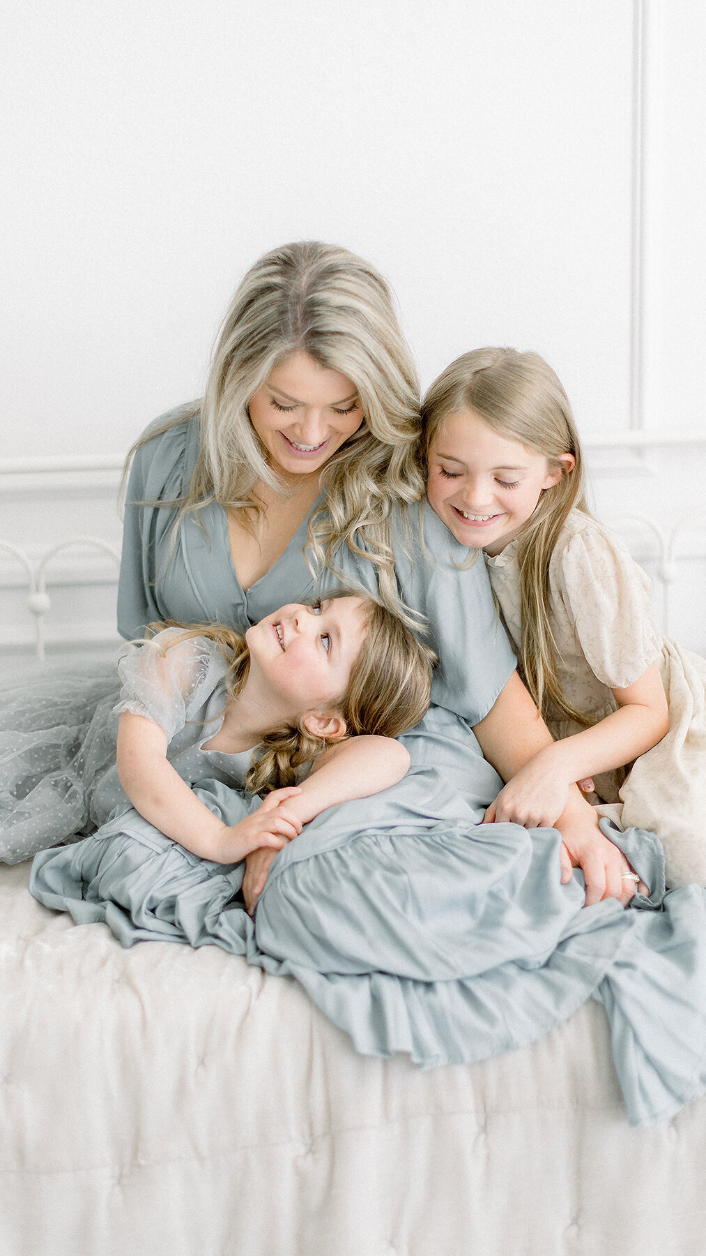 A mother and her two girls in a photography studio cuddling each other while they are sitting on a bed for their family photos together.