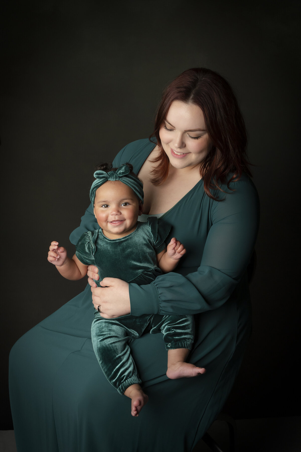 Mom and her eight-month-old daughter in a studio setting, creating a harmonious green color palette with their dress, baby romper and the background. Captured by Charlotte child photographer, Alicia Insley Smith. Capture precious memories with your little one in a timeless motherhood portrait