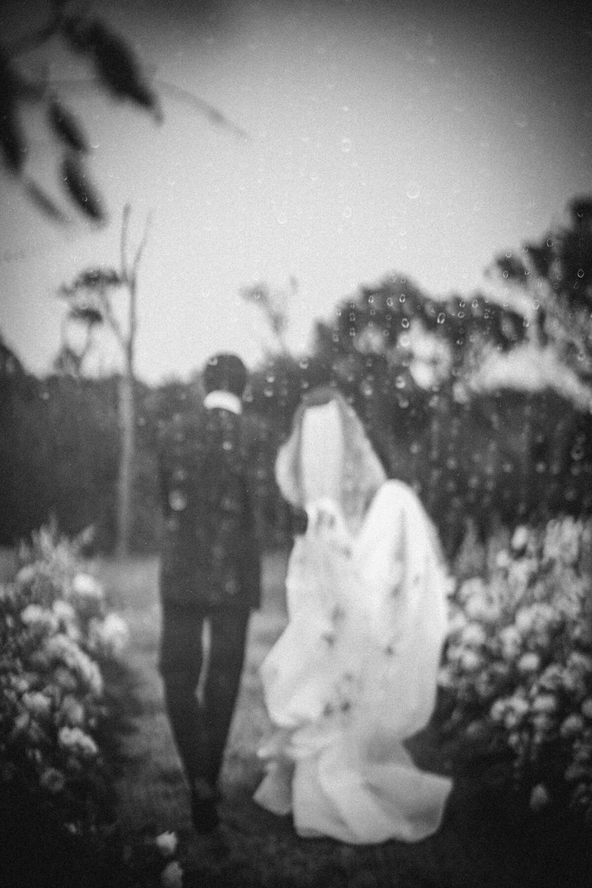 A couple walks outdoors in wedding attire, seen from behind, with raindrops visible in the foreground. The image is black and white, creating a nostalgic atmosphere. This photo was taken by a NYC wedding photographer for vintage-style weddings in Brooklyn