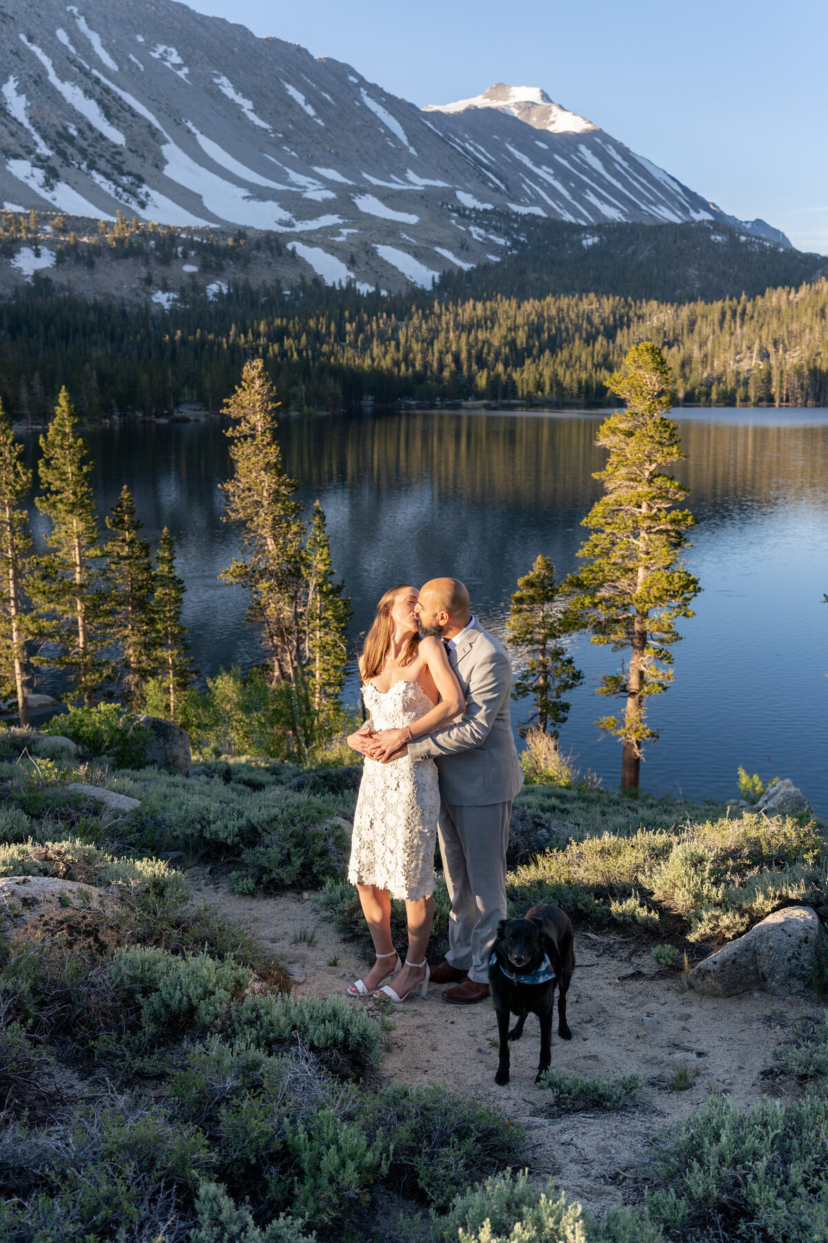 A newlywed couple kissing in front of a lake with a dog in front of them
