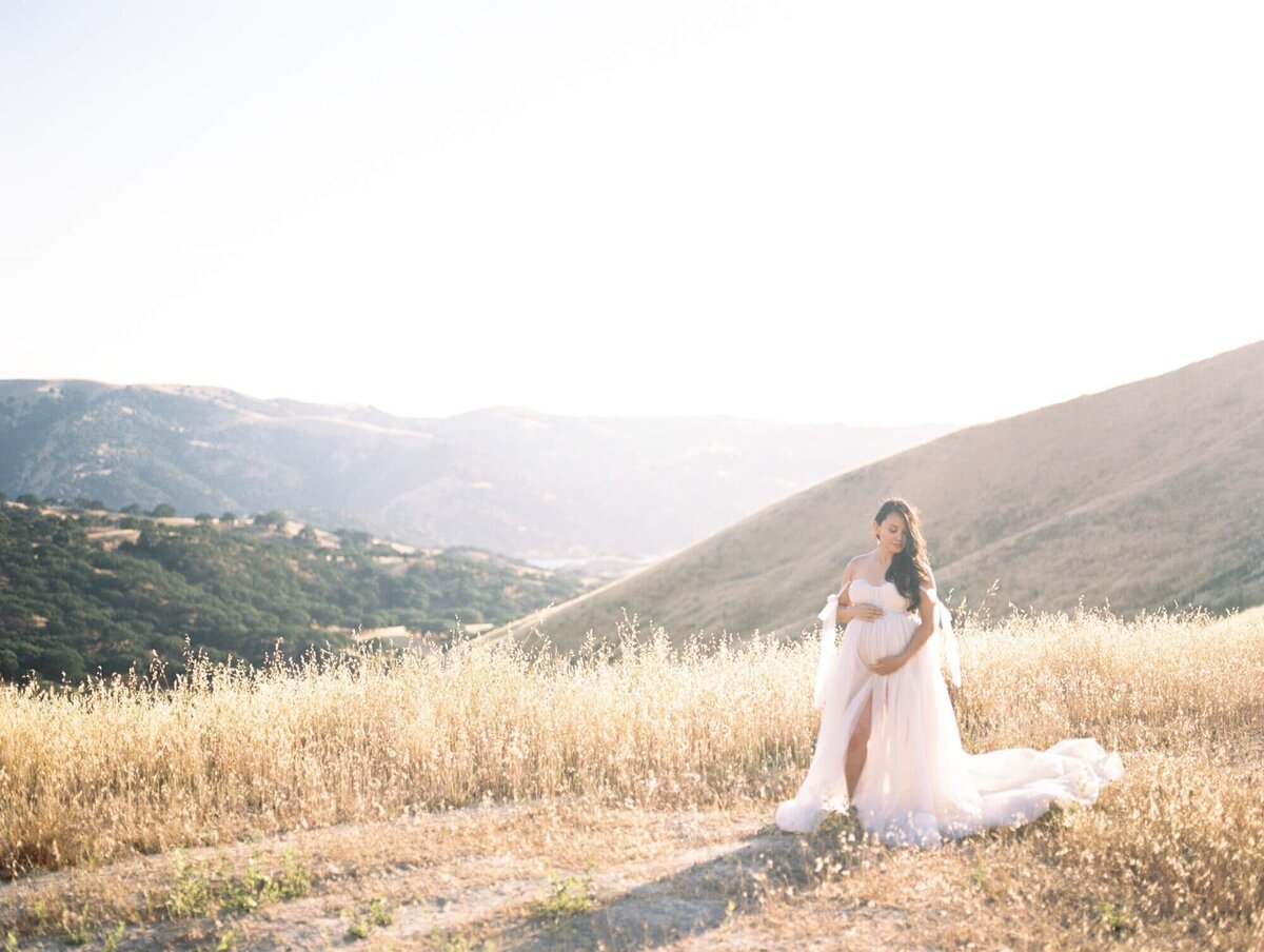 Hillside maternity photoshoot in San Francisco with a pregnant woman standing in a field against hills.