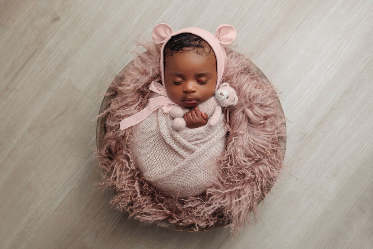 Newborn baby wrapped in a soft pink blanket, wearing a matching bear hat, sleeping in a round wooden bowl lined with fluffy pink fur, posed on a light wooden floor.