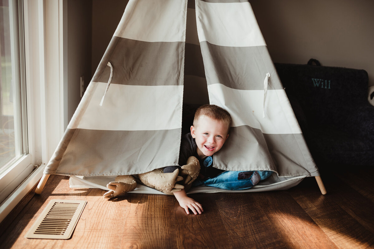 A little boy is peeking out of the teepee and laughing.