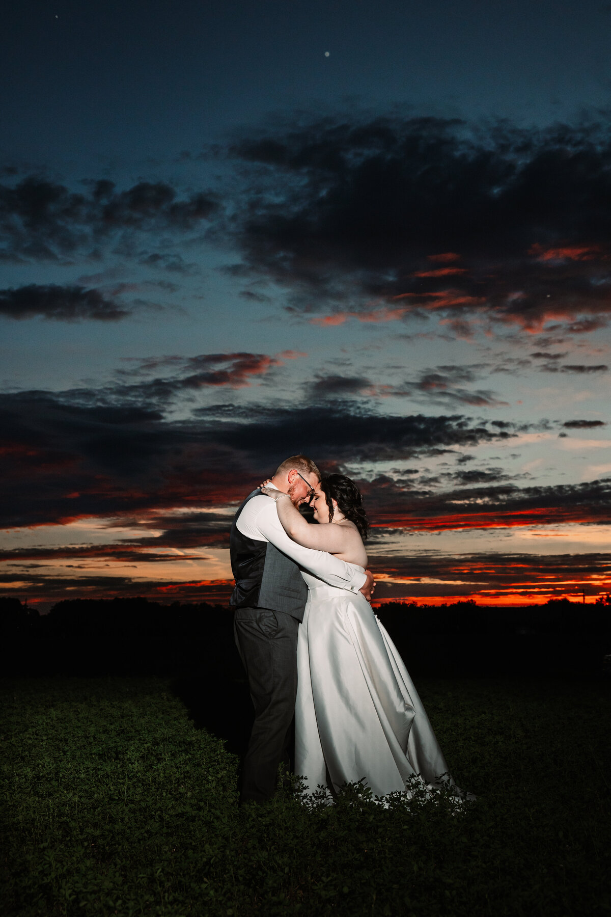 A bride and groom touch foreheads in a field under a red sunset