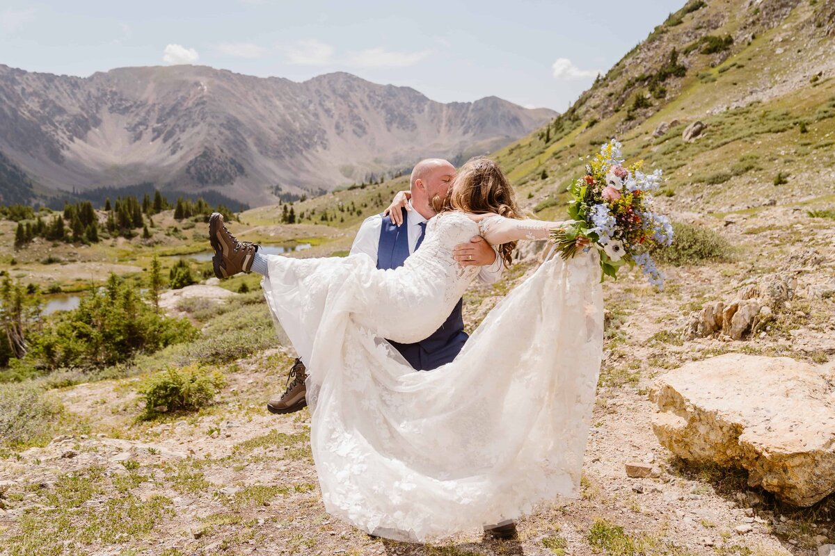 Groom carries bride in his arms while kissing her, surrounded by sweeping mountain views at Loveland Pass in Colorado.
