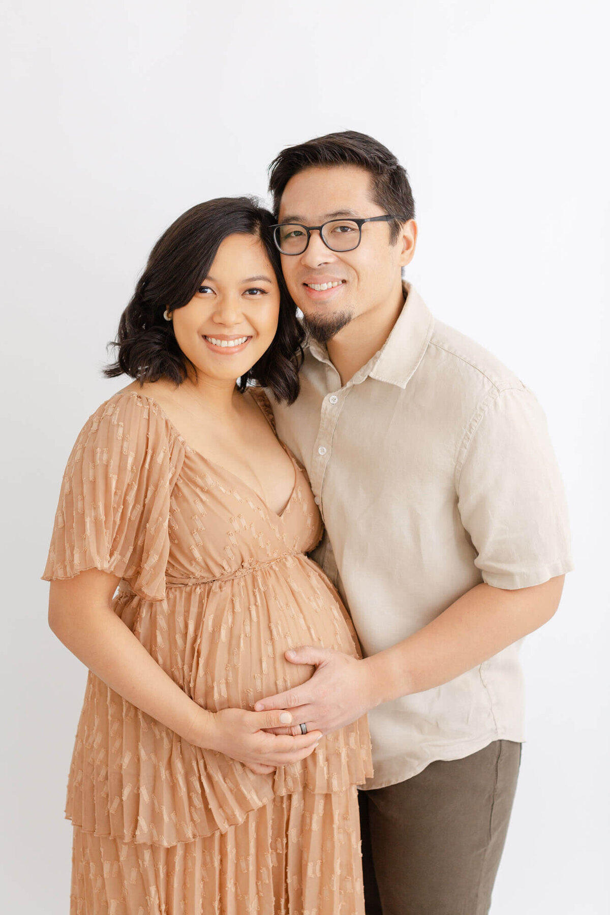 Mama in a taupe dress and standing next to husband who is in neutral clothes. They have their faces touching and are both smiling at the camera while holding Mama's baby belly.