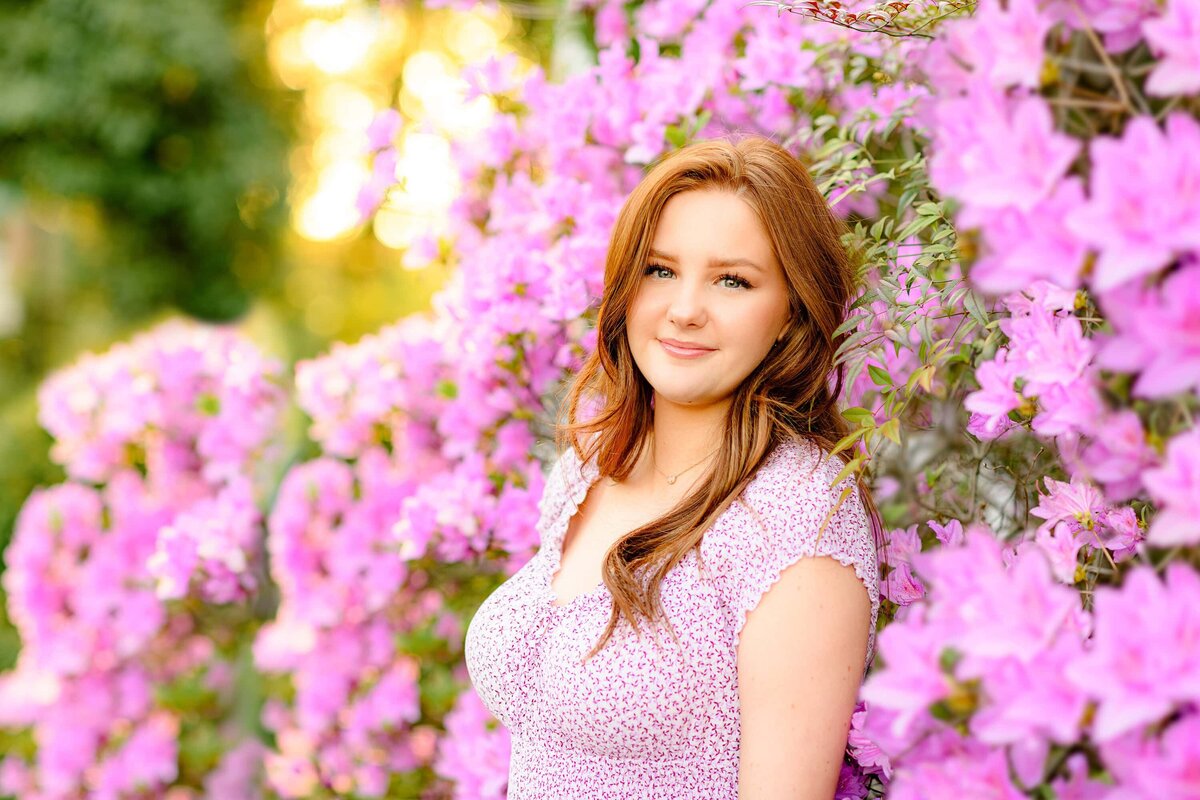 red-headed Charlotte area senior dressed in a purple dress stands with her back to a azelea bush with purple blooms in glen cairn gardens in sc