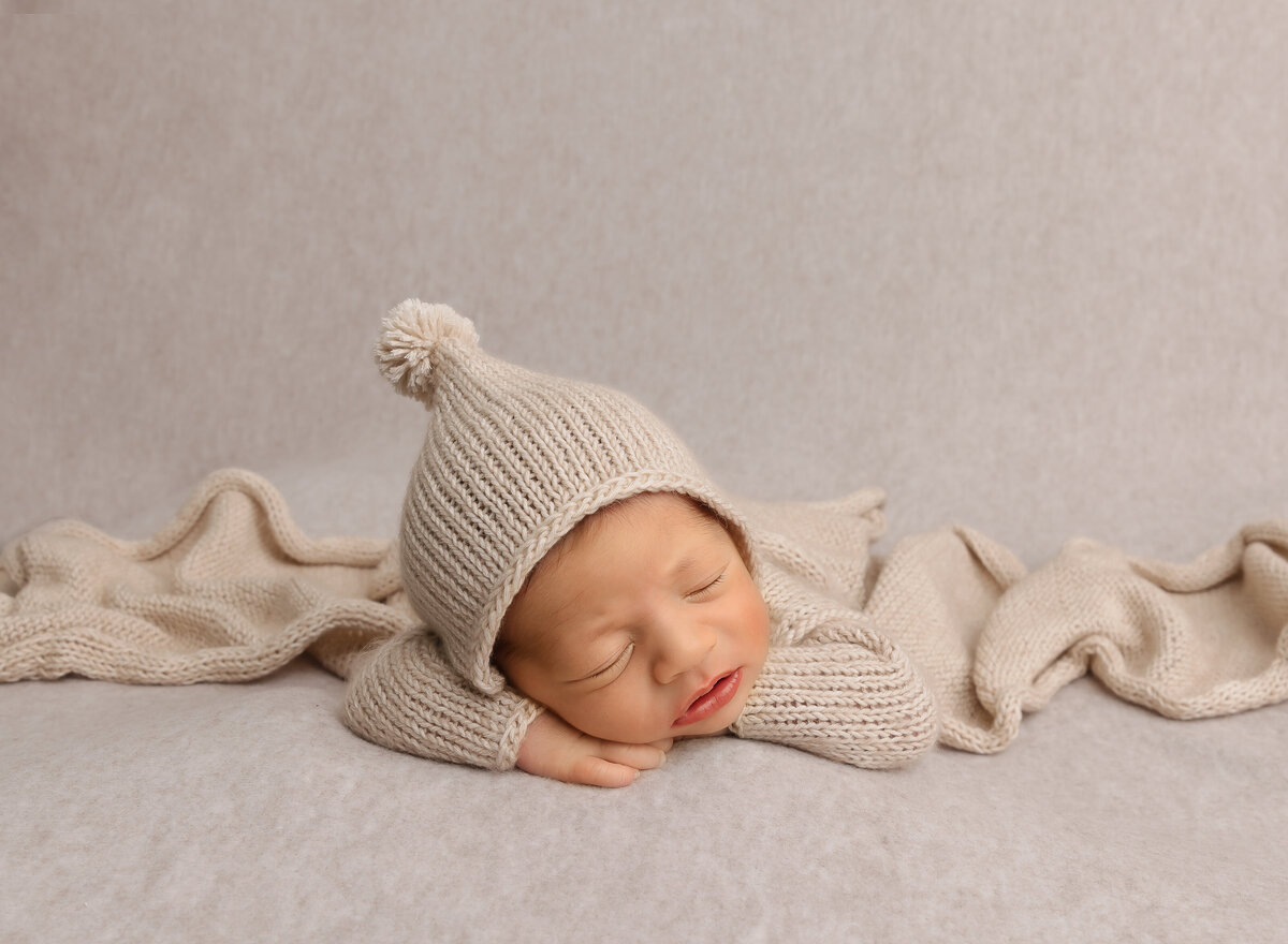 Brooklyn newborn photoshoot. Newborn baby is sleeping on his belly with his hands folded under his cheek. Baby is wearing a beige knit onesie and cap.  Captured by premier Brooklyn NY newborn photographer Chaya Bornstein Photography.