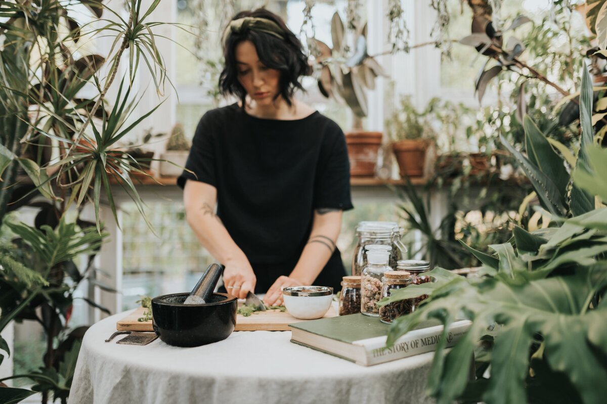 Woman stands in lush, greenhouse wearing a headband and black t-shirt while chopping herbs on cutting board atop a table with linen tablecloth.