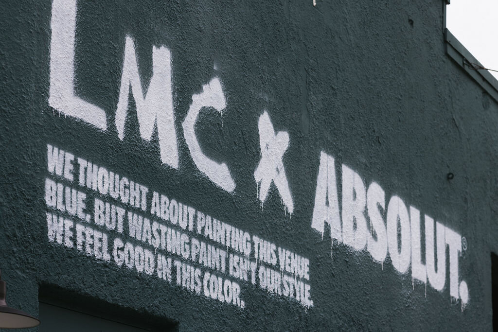 Exterior wall with white stencil text “LMC & Absolut” and slogan “We thought about painting this venue blue, but wasting paint felt unwise. We feel good in this color” at the RDEE-produced LMC x Absolut activation at Tales of the Cocktail 2025.