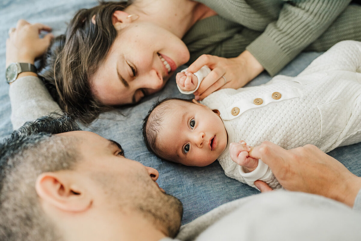 new parents lay on bed snuggling while newborn stares at camera