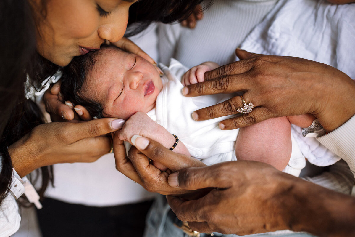 mom leaning over and kissing sleeping newborn held by grandparents
