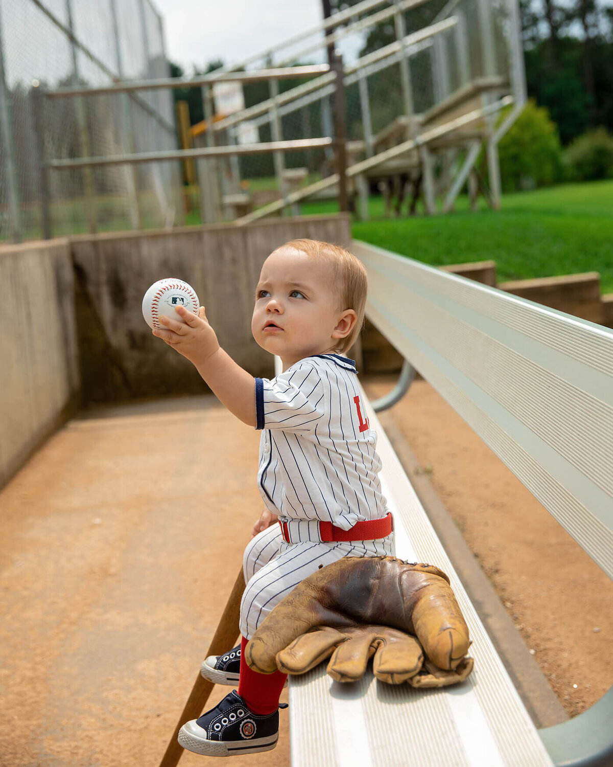 1 year old boy sitting on the dugout bench holding a baseball.