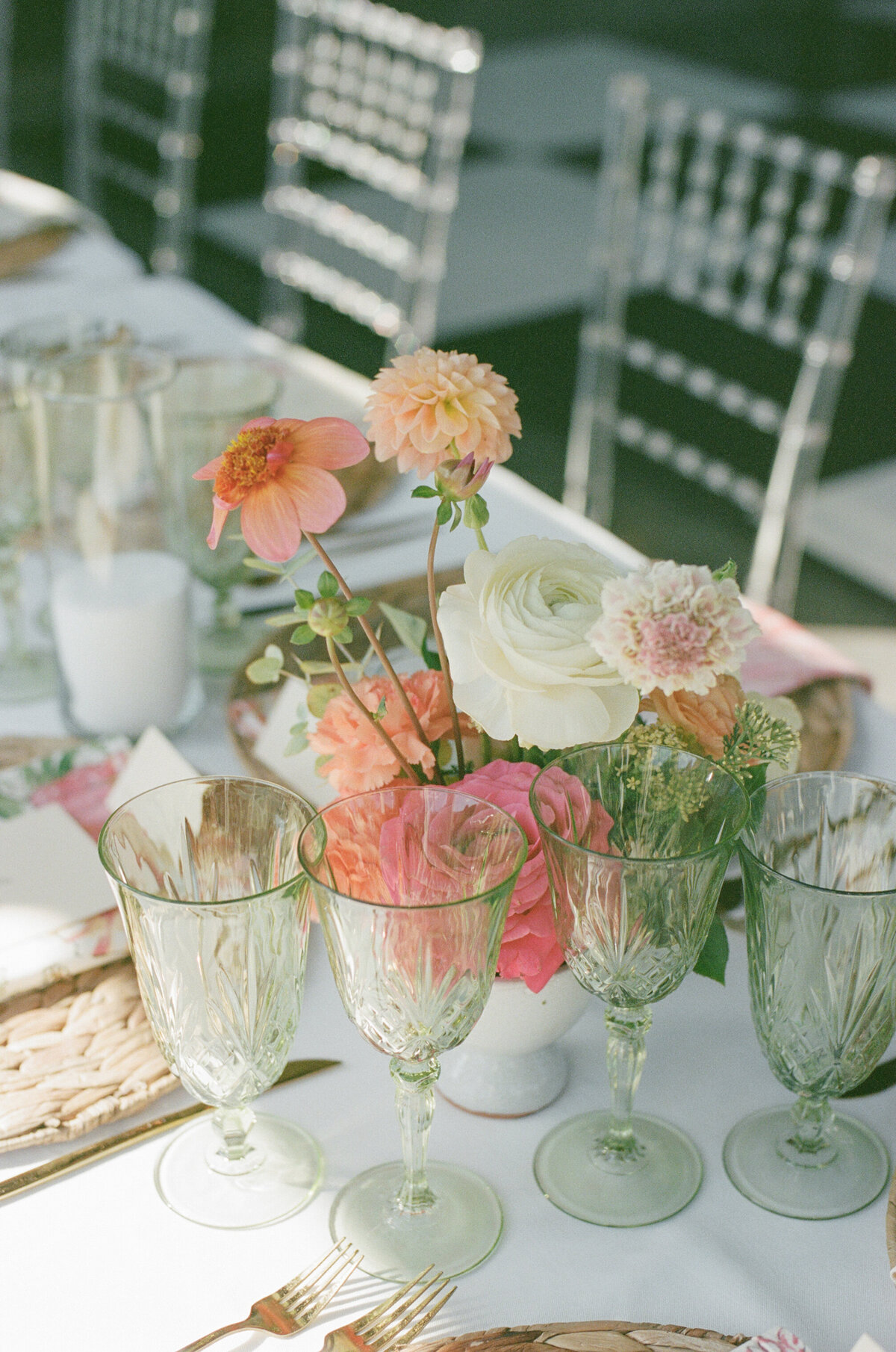 Wedding reception table decorated with a delicate peach and pink floral centerpiece featuring dahlias, ranunculus, and roses, paired with vintage-inspired glassware for an elegant garden party aesthetic.

