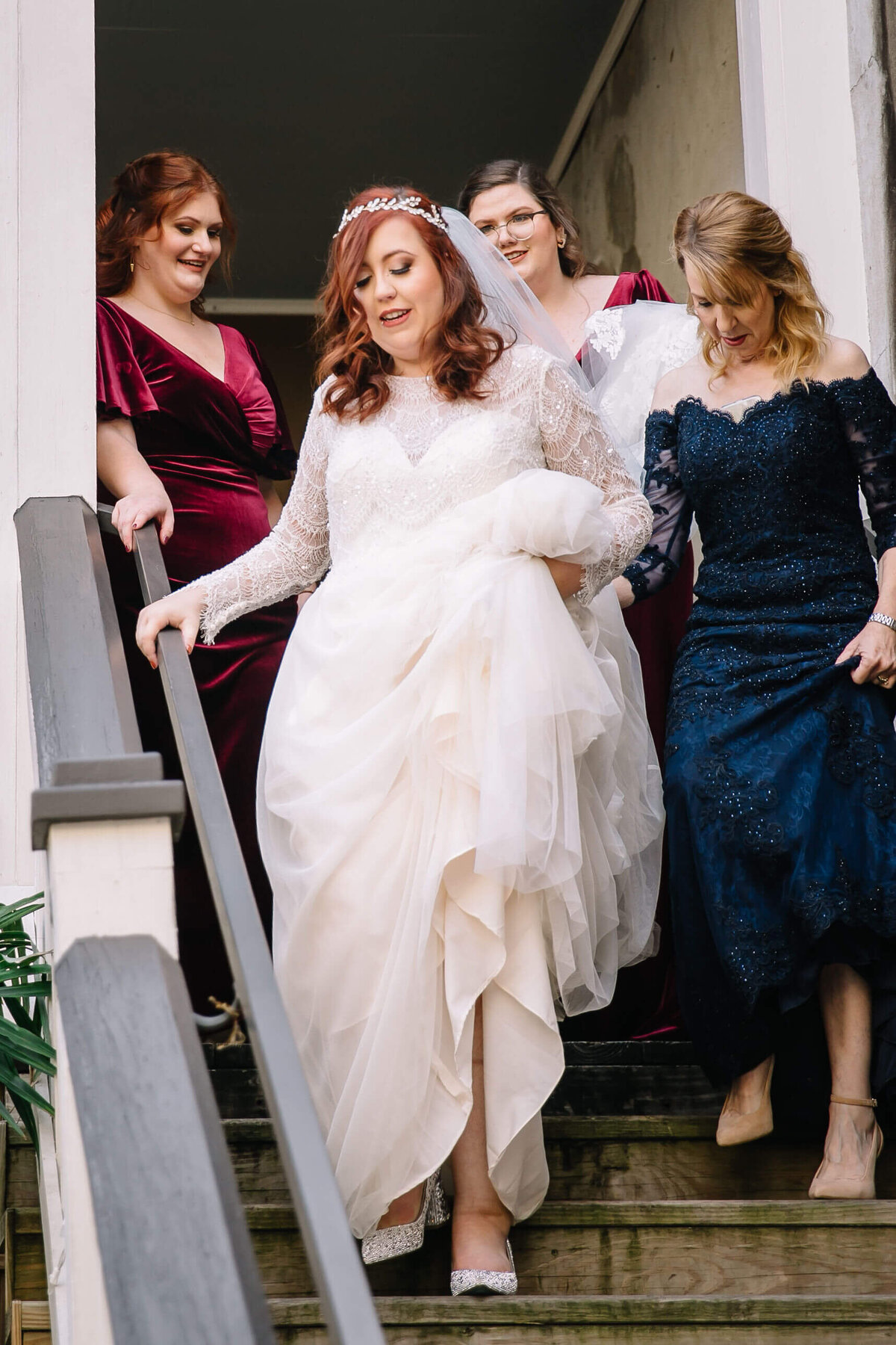 A bride in a white dress and veil walks down wooden stairs, smiling, assisted by three women in elegant dresses, two in burgundy and one in navy blue.