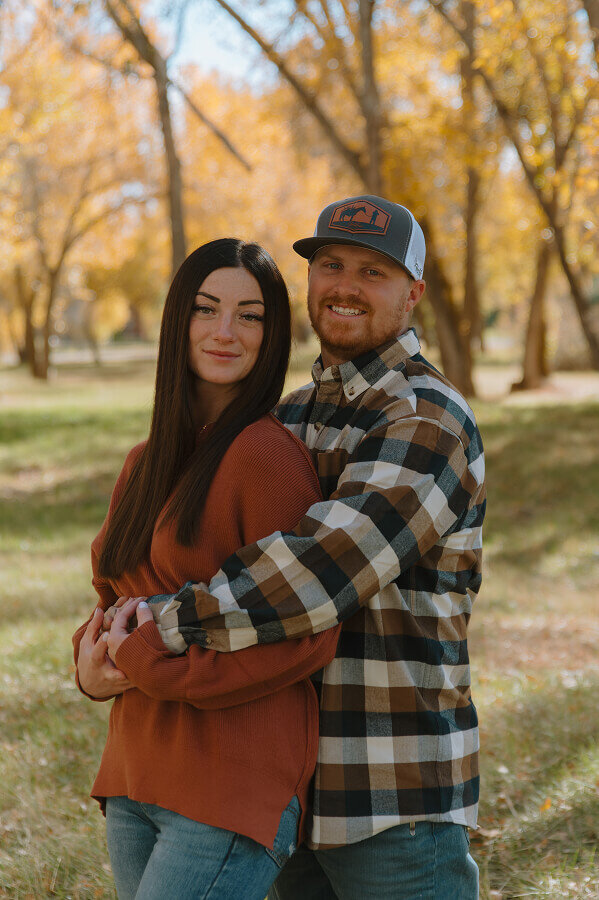 A husband holds his wife from behind as their smile for a photo. She's holding his hands in front of them.