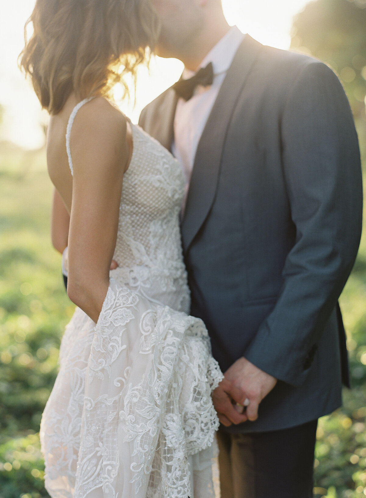 A romantic Caribbean destination wedding scene captured by photographer Laura Gordon. A beautiful bride in a lace wedding dress and her groom in a suit and bow tie, stand closely together in the Dominican Republic. The groom holds the bride's hand as they gaze at each other, bathed in soft sunlight, creating a dreamy tropical ambiance.