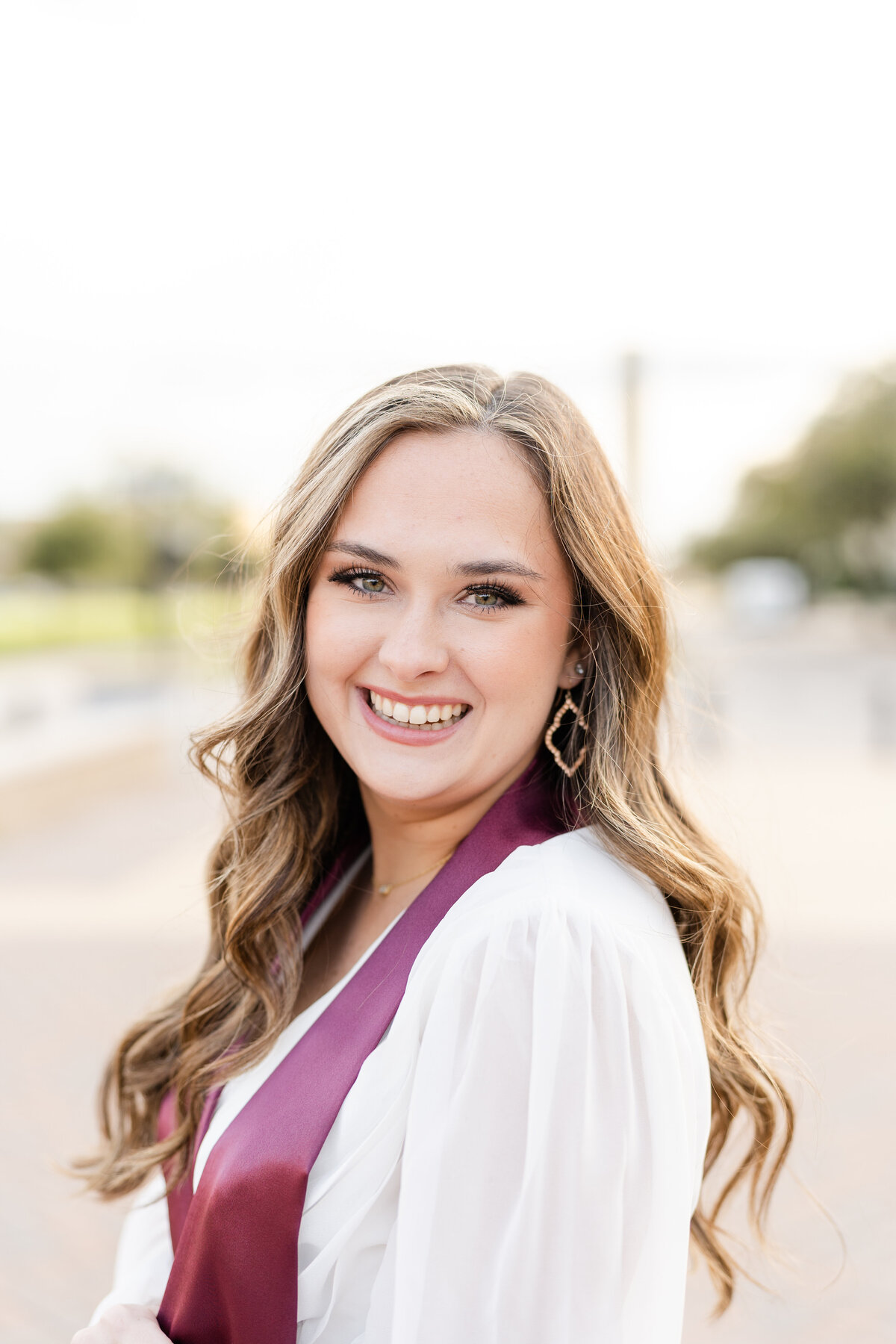 Texas A&M senior girl smiling over shoulder while wearing white dress and maroon stole in front of Bell Tower at Military Plaza