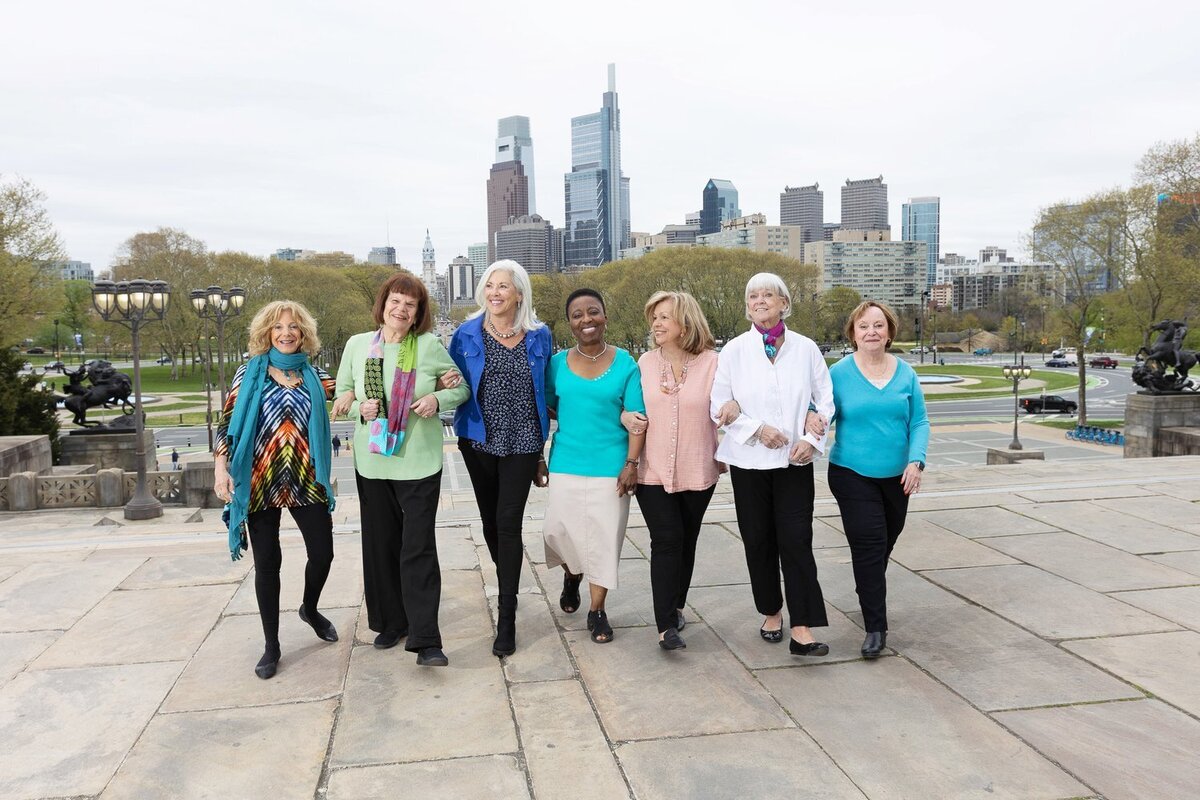 7 older women dressed casually walking towards the camera with the Philadelphia skyline behind them