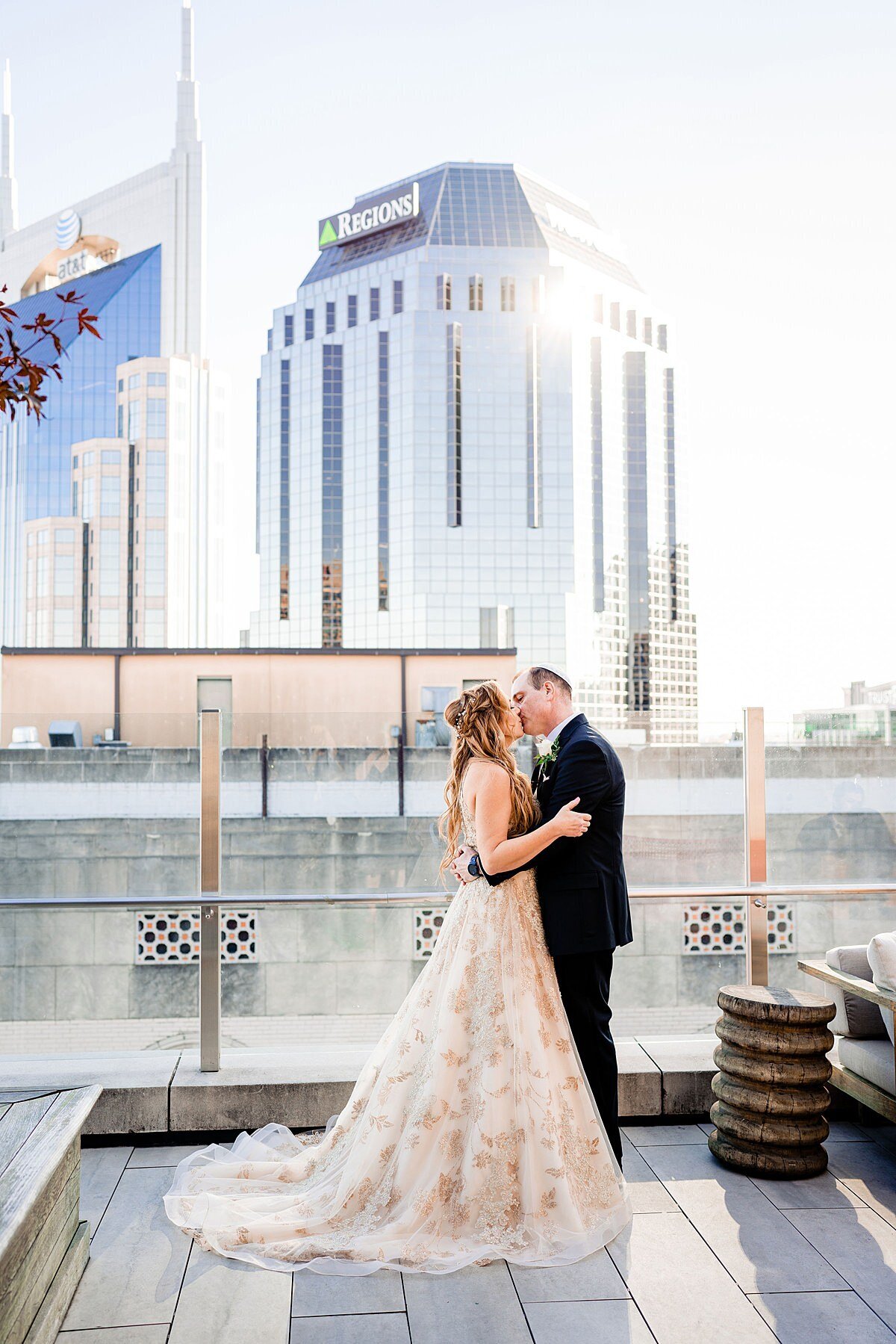 Jewish bride wearing a gold wedding gown and Jewish groom wearing a black tuxedo kiss on the rooftop of the Noelle Hotel in Nashville, TN with the Nashville Skyline in the background