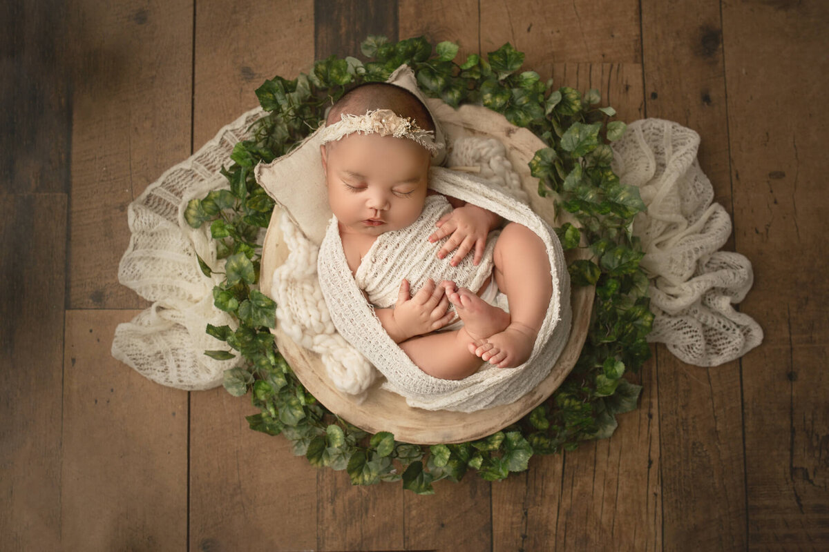 hamilton newborn photographer captures new baby wrapped in white, surrounded by greenery and placed on a dark wood backdrop