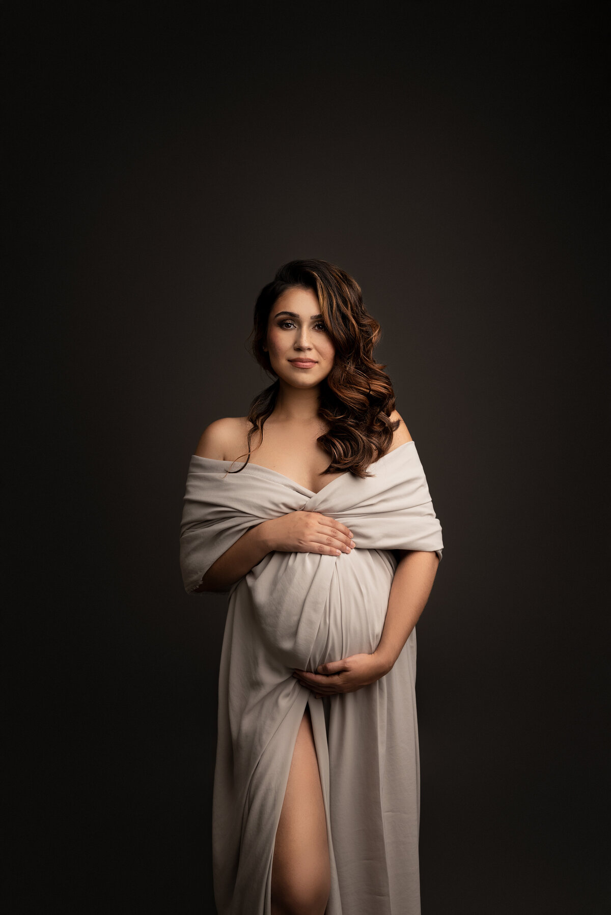 In this captivating photograph, Katie Marshall, the renowned Main Line maternity photographer, skillfully captures the essence of an expectant mom. The radiant mother stands gracefully, facing the camera, donned in an elegant linen-colored off-the-shoulder dress. Her flowing reddish-brown curls cascade gently over her shoulder, framing her serene expression as she gazes directly into the camera, her smile exuding warmth and anticipation.