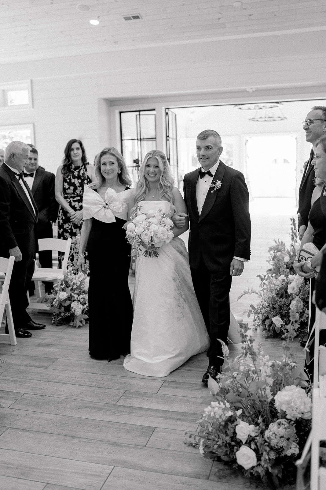 Bride walking down the aisle with parents at indoor ceremony surrounded by florals.