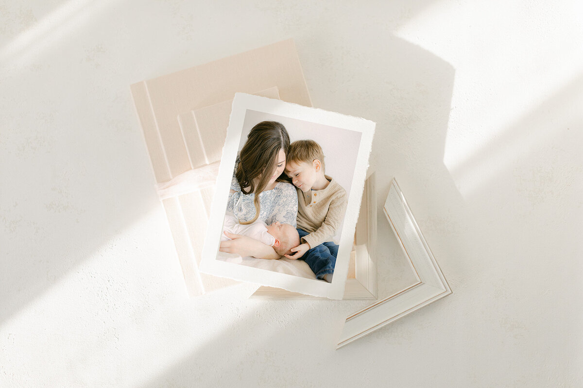 Flat lay of heirloom albums, a white frame corner, and a fine art image of a mother, her toddler son, and her newborn baby boy, products offered by Indianapolis newborn photographer, Brittney Lear.
