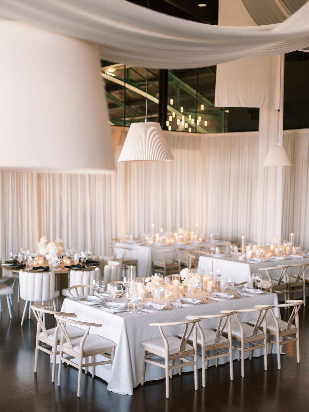 A wedding reception with white drapery, hanging modern lamps, and white tables set with elegant floral arrangements.
