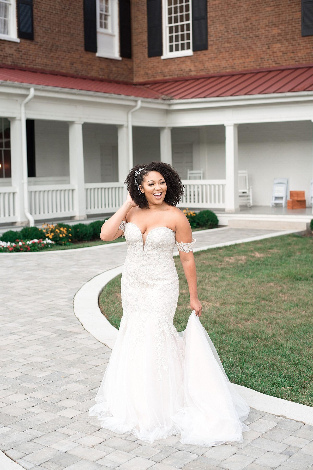 The African America bride, Jasmine Sweet, wearing an off the shoulder lace mermaid wedding gown with a long train smiles at her wedding guests as she walks down the path at the back of Ravenswood Mansion towards the courtyard where her guests are waiting.