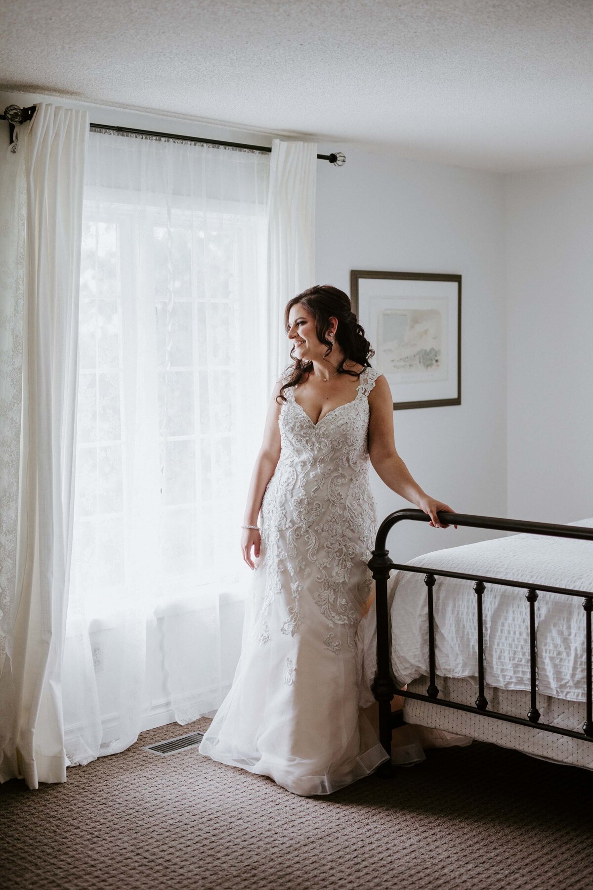 Bride is posing for a photo in the bridal suite before her wedding. She has one hand gently resting on the foot of the bed while looking out the window.