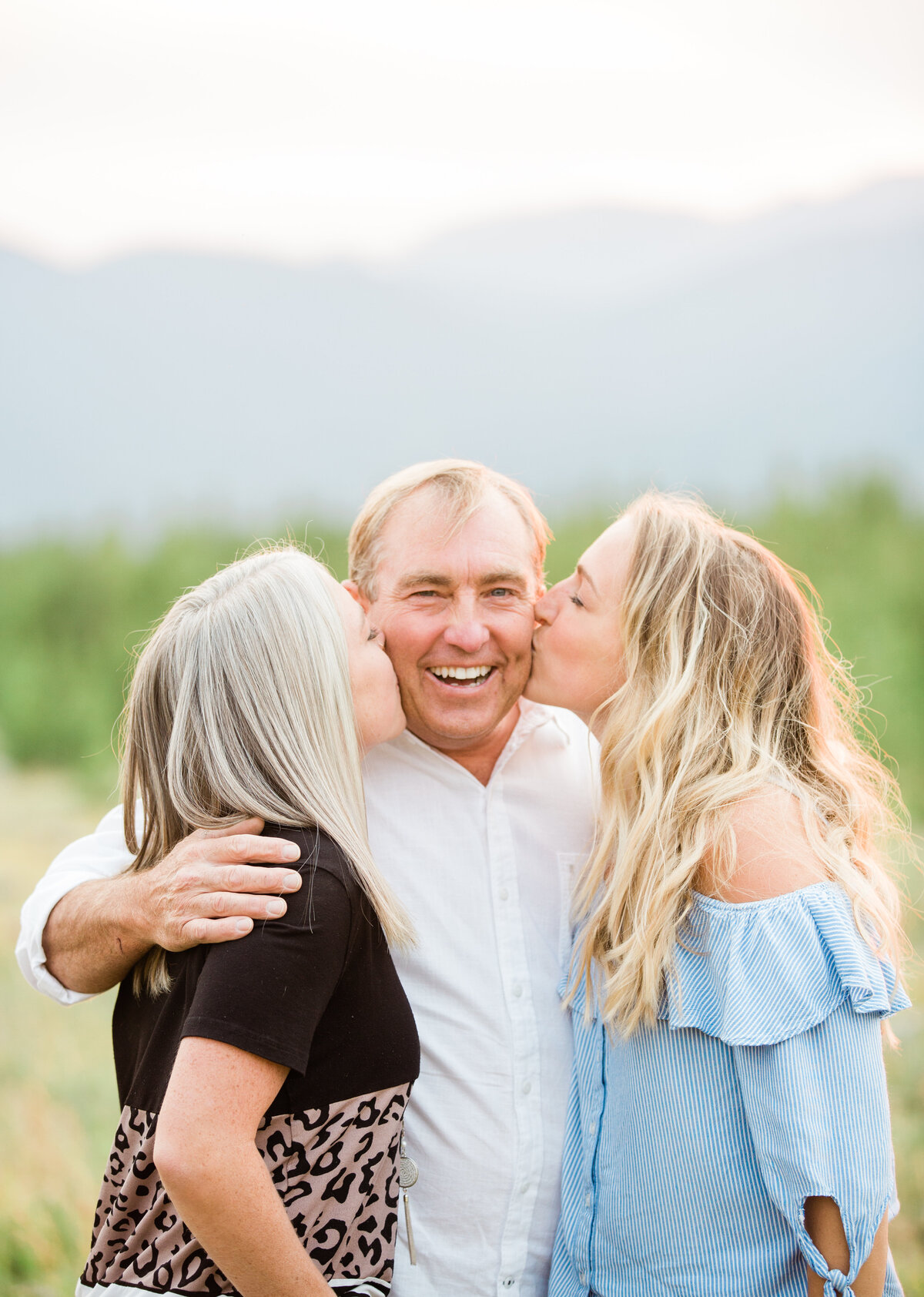 A dad is receiving a kiss on the cheek by each of his adult daughters