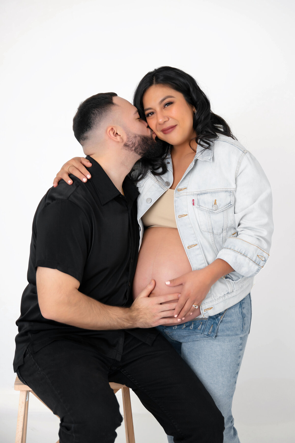 Expecting parents touch mom’s belly as dad kisses her cheek in an intimate moment.