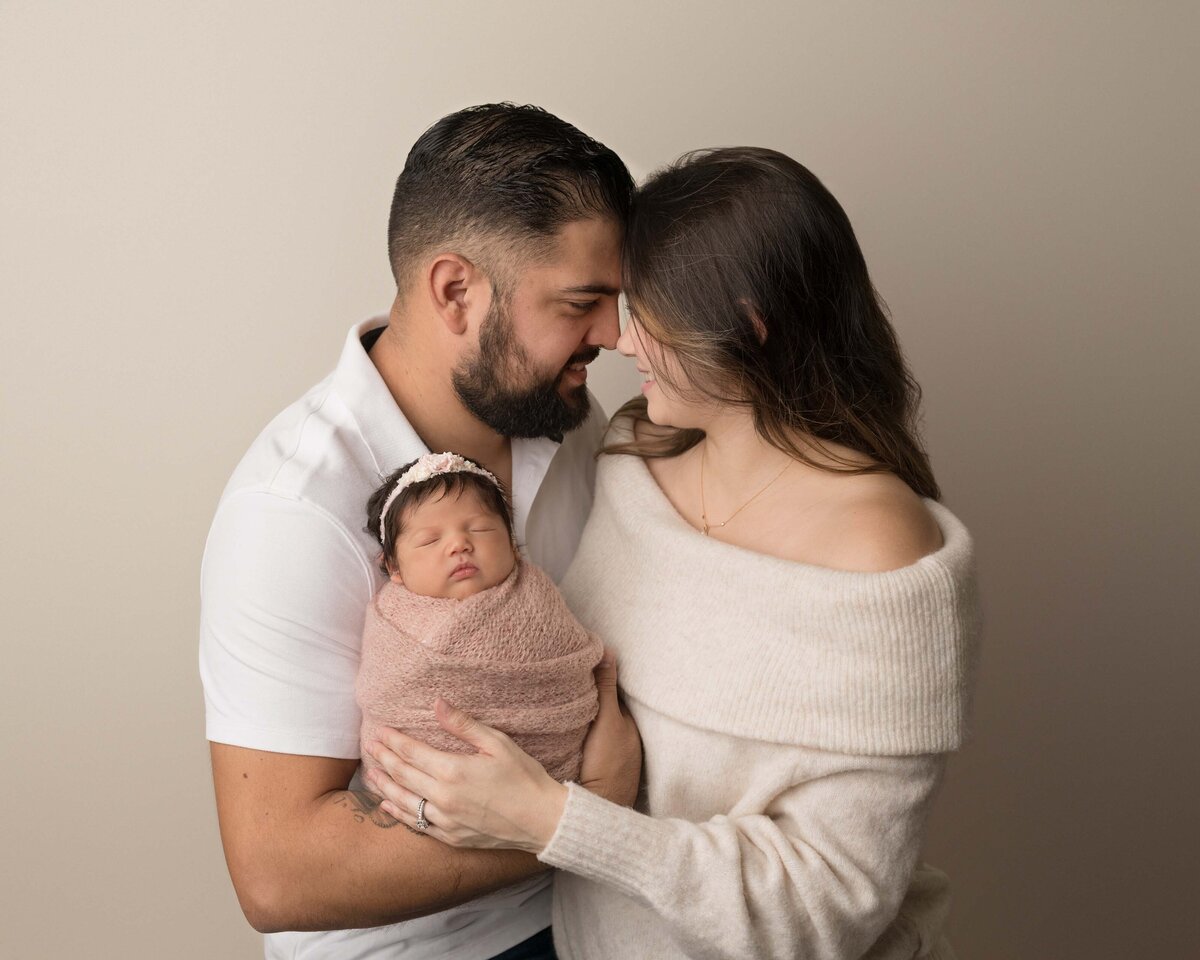 Mom and Dad pose for a newborn photoshoot with their newborn baby girl. Dad is holding baby so she is facing the camera, mom has her hand resting on the baby. Mom and dad are touching their foreheads and smiling. Captured by best Lake Elsinore newborn photographer Bonny Lynn Photography