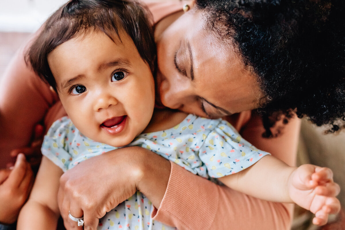 A close-up of a smiling baby being held by their mother, who is gently nuzzling the baby's cheek.