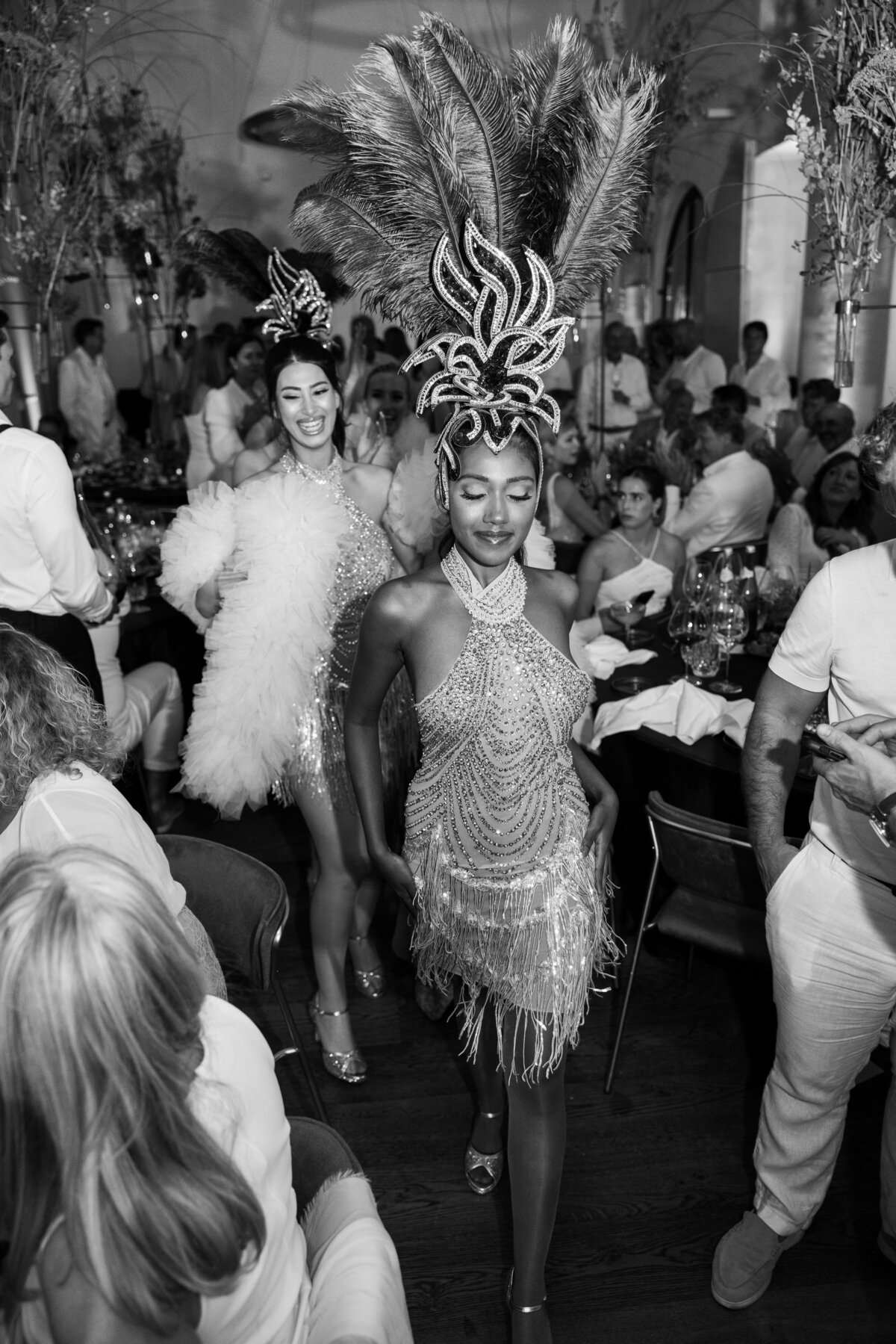 Two exotic dancers in sparkling costumes lead a parade through a banquet hall full of guests. The black-and-white image highlights the elegance and vibrancy of this exclusive event.