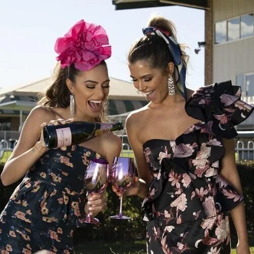 Stylish women in floral race day outfits popping champagne and toasting with branded Melbourne Cup Carnival glasses