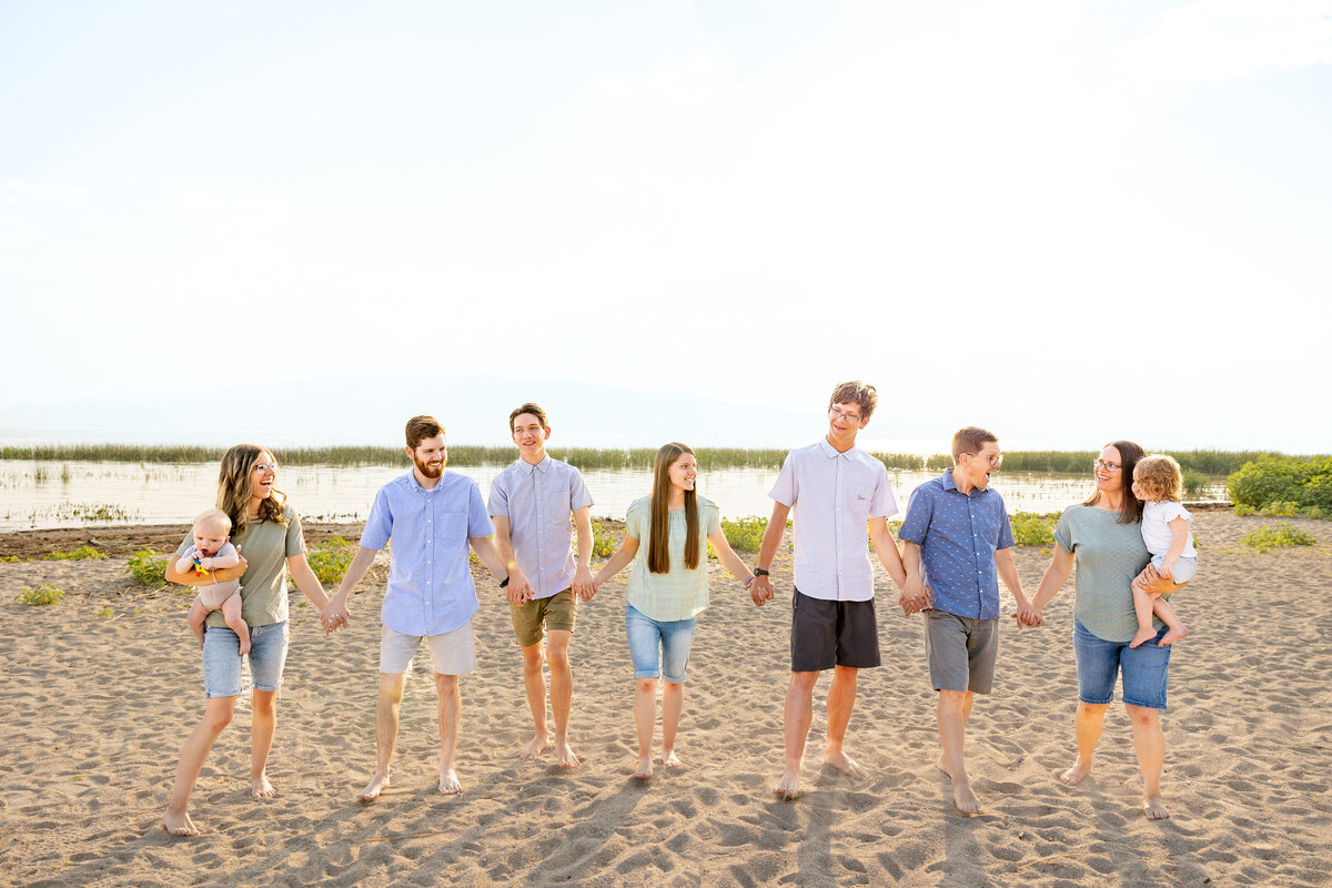 A family walks hand in hand on a sandy beach, enjoying a relaxed and loving moment. The clear sky and peaceful waters create a serene backdrop as they bond in the golden hour. Taken by Cassie Lee Photography in Vinyard Utah