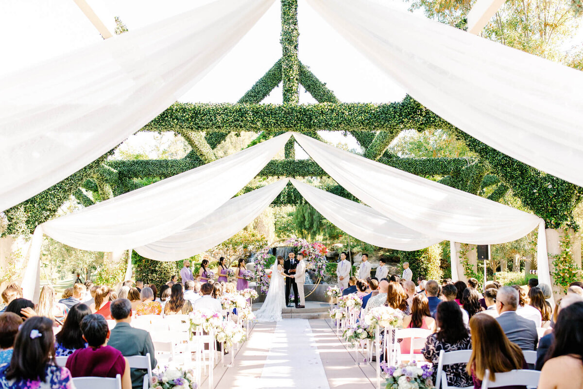 Coyote Hills Golf Course wedding ceremony under a pergola adorned with flowing white drapes and greenery. Guests are seated on either side, facing the couple and officiant. Bright, joyful atmosphere.
