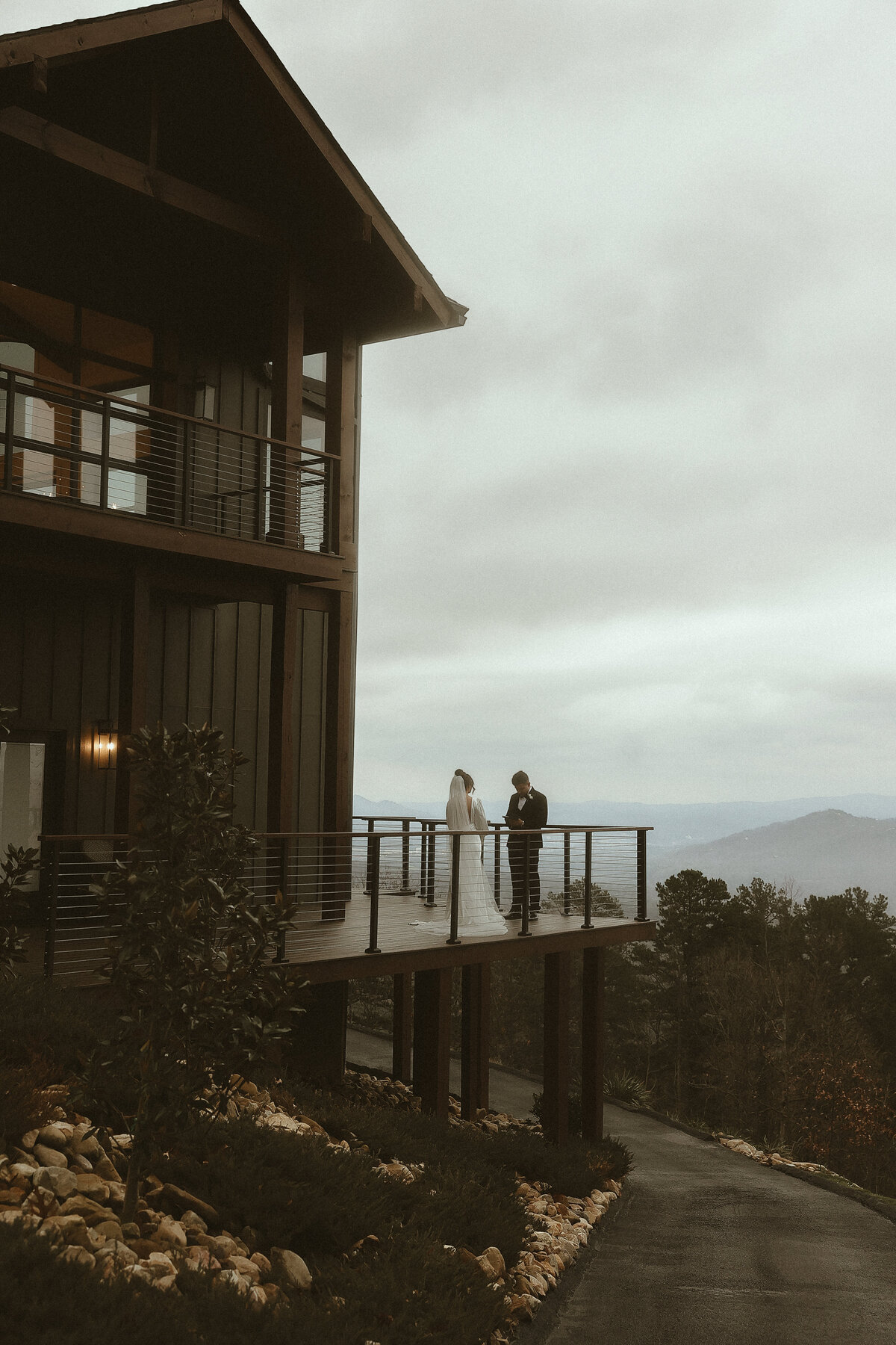 A couple stands on a scenic balcony, exchanging vows with a mountain view under a cloudy sky, framed by a modern, wooded house.